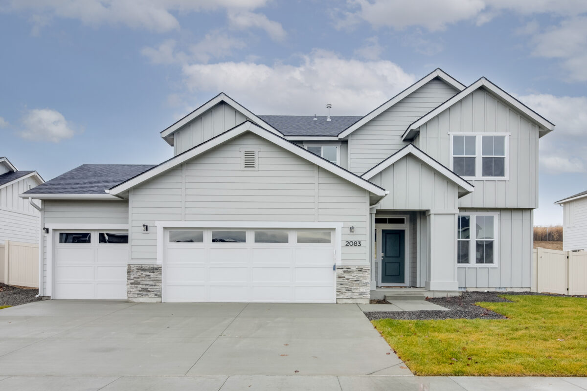 Front view of a modern two-story home in a CBH Homes community, featuring light gray siding, a dark gray roof, white garage doors, stone accents, and a landscaped front yard, perfect for homebuyers in Idaho.