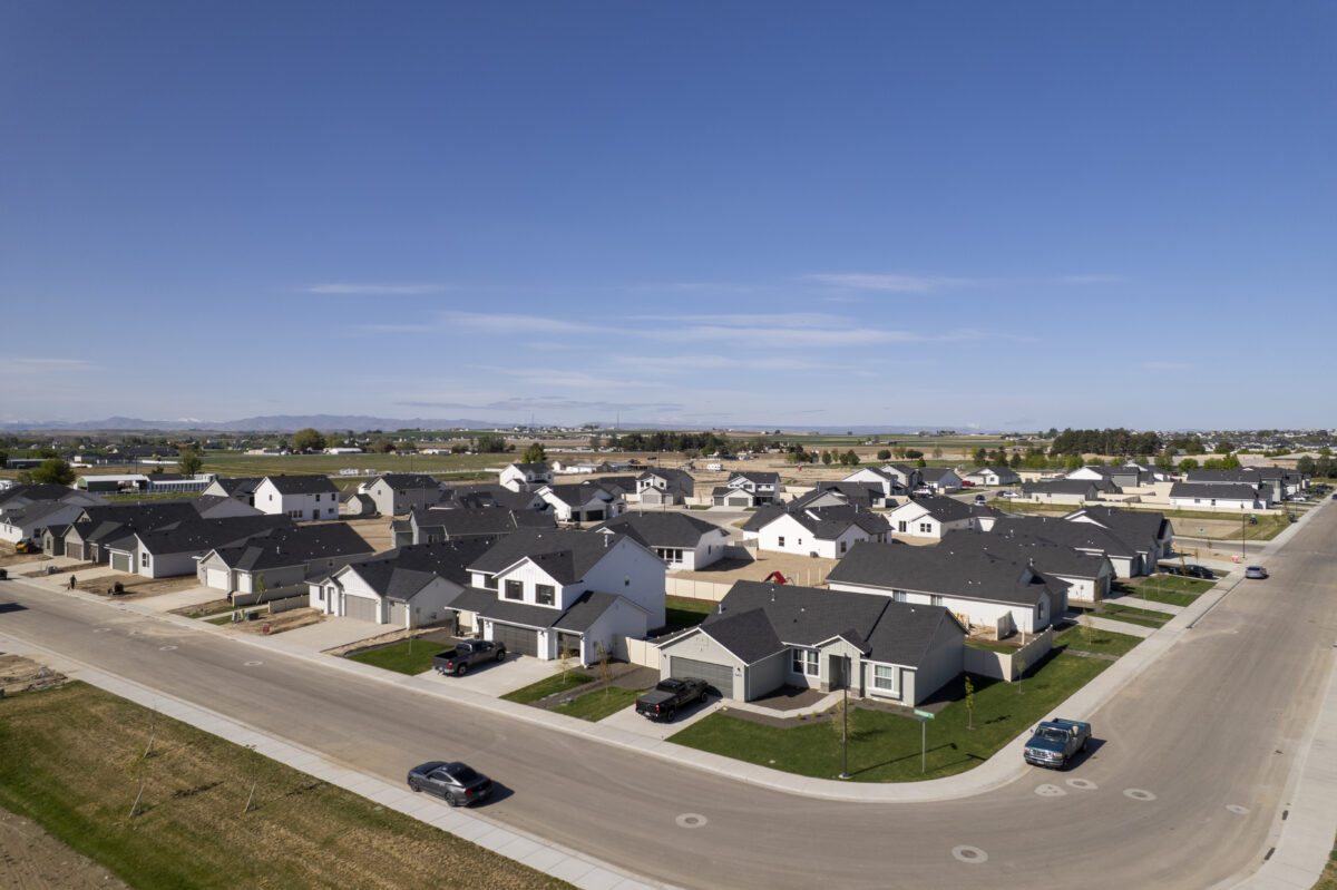 Aerial view of a suburban neighborhood with a mix of single-story and two-story modern homes arranged along wide, newly paved streets. Some homes are fully completed while others are under construction. The development is surrounded by expansive farmland with distant hills and a clear blue sky overhead, suggesting a peaceful rural setting.