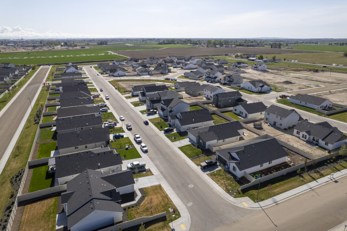 Aerial view of a suburban housing development bordered by open farmland and a narrow irrigation canal. The neighborhood features curving streets lined with modern two-story and single-story homes, some with landscaped yards and others still under construction. The backdrop includes wide agricultural fields, clusters of trees, and distant hills under a clear sky with scattered clouds.