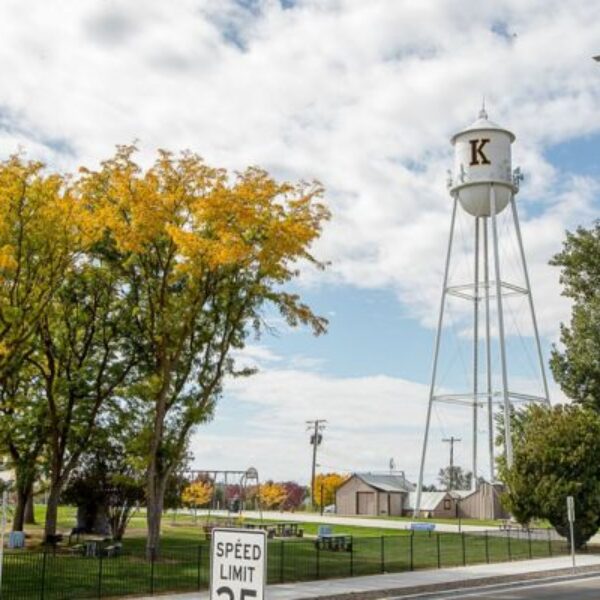 A white water tower with a large "K" painted on its tank stands above a small-town park, with autumn-colored trees, a playground, a speed limit 25 sign, and a residential street in the foreground under a partly cloudy blue sky.