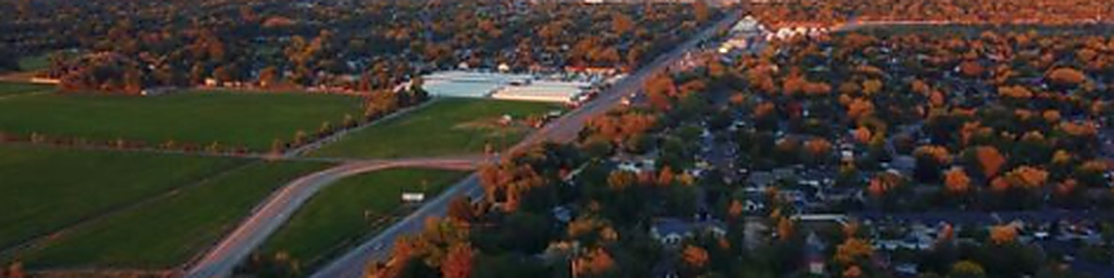 Aerial view of a Boise-area neighborhood at sunset, with tree-lined streets in fall colors, a road cutting diagonally through the scene, and green fields in the foreground.