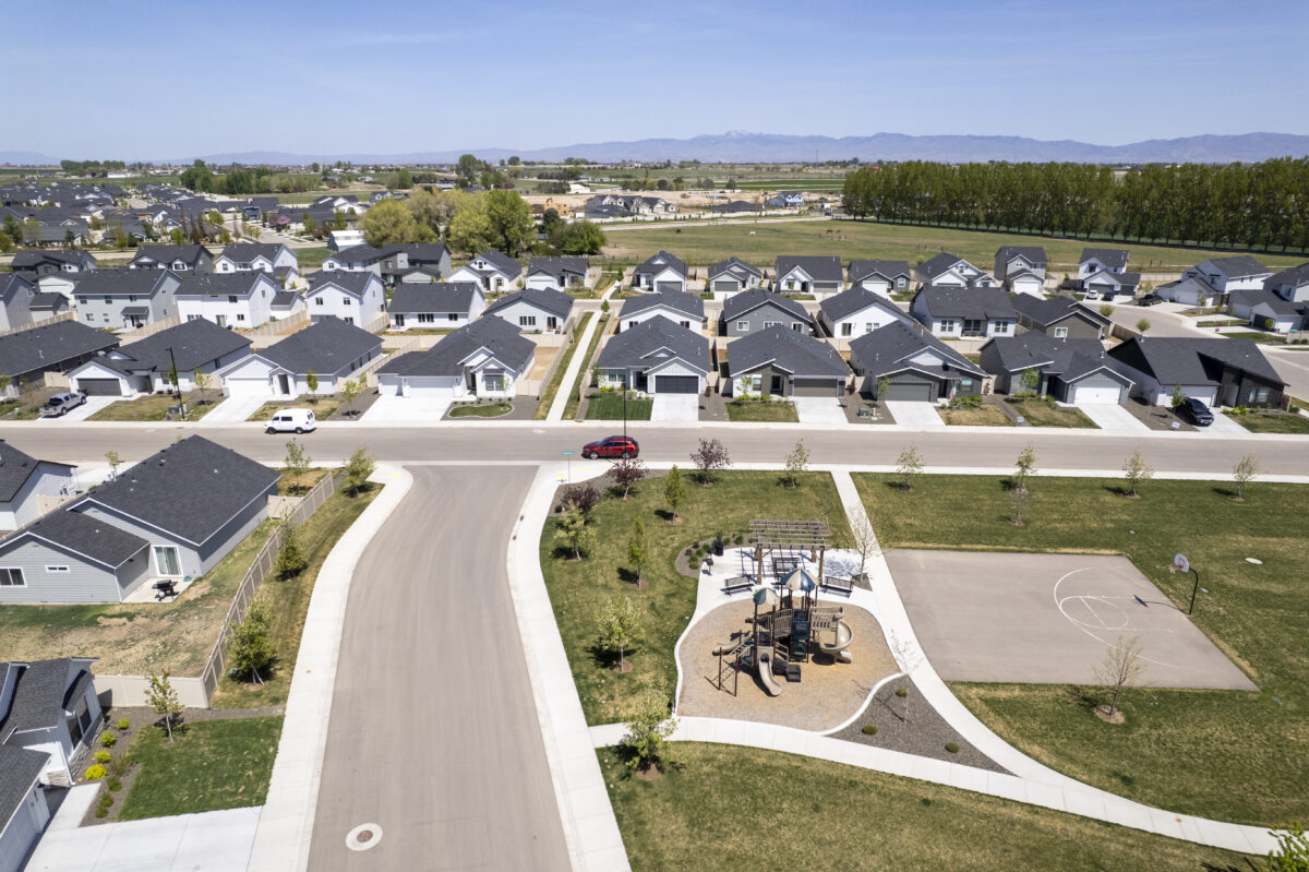 Overhead view of a suburban neighborhood park nestled within a residential development. The park includes a playground with slides and climbing structures, a half basketball court, and walking paths surrounded by open grassy areas and young trees. Rows of single-family homes with garages and driveways line the streets, and farmland and trees stretch into the distance under a clear blue sky.