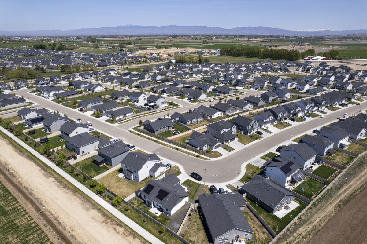 Aerial photo of a large, neatly arranged suburban neighborhood surrounded by farmland. The development features wide, curving streets lined with single-family homes, most with fenced backyards and paved driveways. A concrete path runs along the outer edge of the neighborhood. In the distance, green fields stretch toward a range of mountains beneath a clear blue sky.