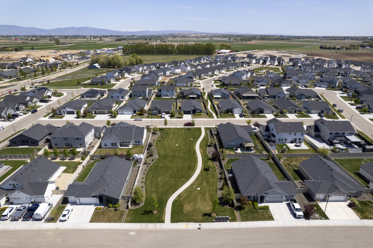 Aerial view of a modern suburban neighborhood featuring rows of single-family homes with gray and black rooftops, landscaped yards, and paved driveways. A curved walking path runs through a central green space lined with young trees. The surrounding area includes open farmland, construction sites, and distant mountain ranges under a clear blue sky.