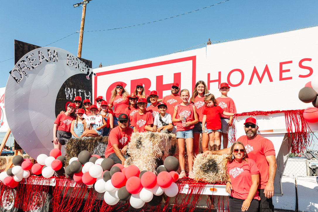 “CBH Homes team and families pose on a decorated float at the 2025 Meridian Dairy Days Parade, wearing matching red ‘Home Grown’ shirts, surrounded by hay bales and red, white, and black balloons in front of a large CBH Homes sign.