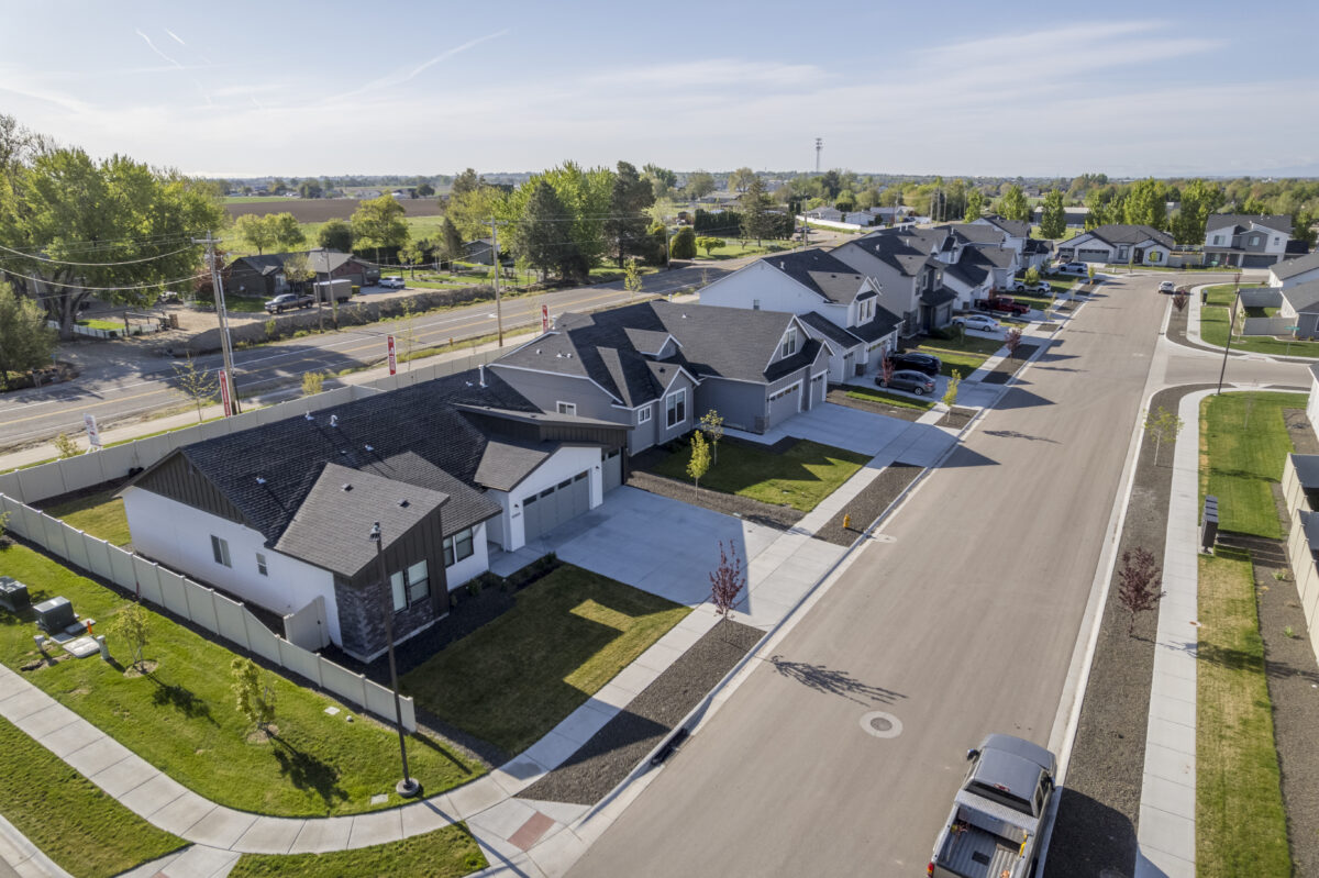 Aerial view of a quiet residential street in a new suburban development. The image shows modern single-family homes with manicured lawns, wide driveways, and a clean, freshly paved road. A row of trees and open farmland can be seen across the main road that borders the neighborhood. The scene is bright and peaceful under a clear blue sky with scattered wispy clouds.