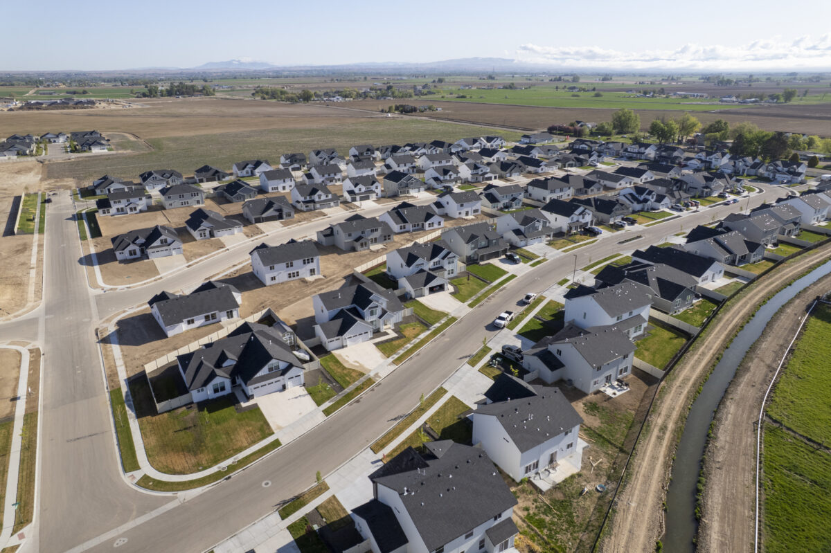 Drone image of a newly built suburban neighborhood with two-story and single-story homes, some on lots still under development. The curved streets are bordered by sidewalks and driveways, and a canal runs along the edge of the community. The neighborhood is surrounded by wide open farmland and distant hills beneath a sunny, clear sky with low-lying clouds on the horizon.