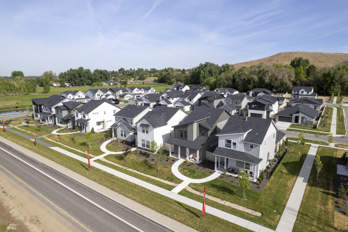 Angled aerial image showcasing new contemporary two-story homes with black and white exteriors along a landscaped main road with red CBH Homes flags, hills and trees in the distance.