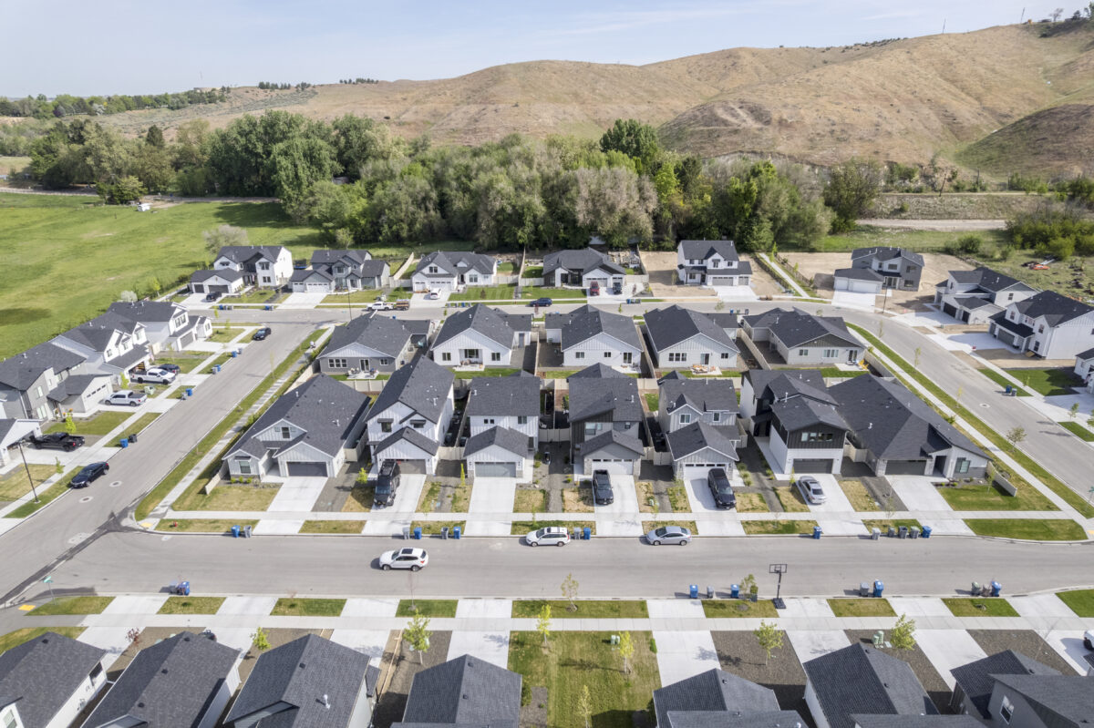 Aerial view of a modern suburban neighborhood with dark-roofed single-family homes arranged in a neat grid pattern, surrounded by green space and foothills in the background.
