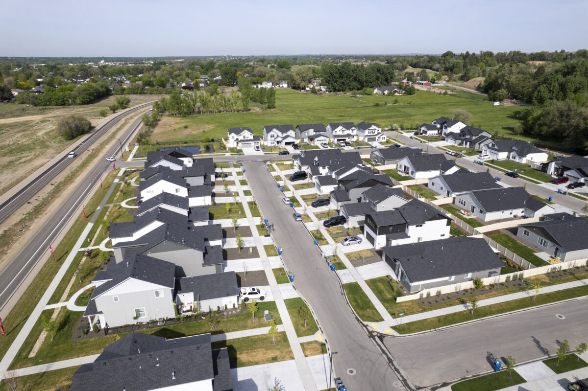 Aerial view of a modern suburban neighborhood with dark-roofed single-family homes arranged in a neat grid pattern, surrounded by green space and foothills in the background.