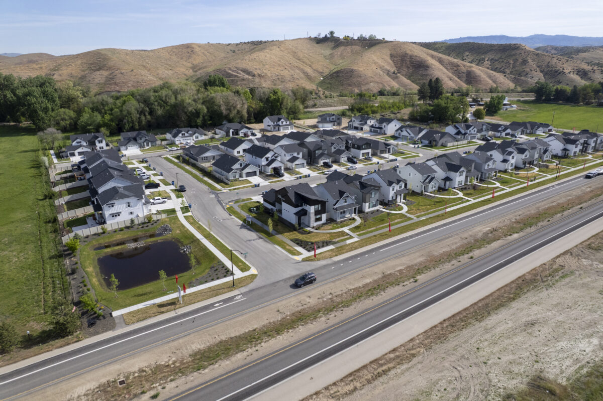 Wide-angle drone view capturing the entire CBH Homes community with its curved front paths, entrance signage, landscaped pond, and scenic rolling foothills.