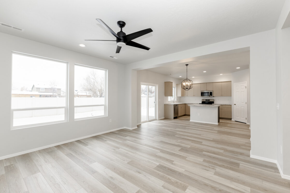 Bright and airy open-concept living area with wide plank light wood-look flooring and large windows offering ample natural light. The space features a modern black ceiling fan, recessed lighting, and a view into the adjacent kitchen and dining area. The kitchen includes natural wood cabinets, a central island with marble-look countertops, stainless steel appliances, and a spherical statement chandelier over the dining nook near sliding glass patio doors.