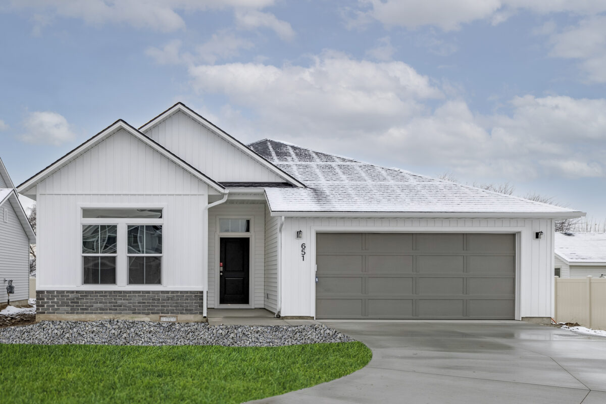 Single-story home with white vertical siding, gray brick accent, and a gray two-car garage. The house has a dark front door with a transom window above and snow lightly dusting the roof. A curved concrete driveway leads up to the entrance, with green grass and decorative rock landscaping in front.