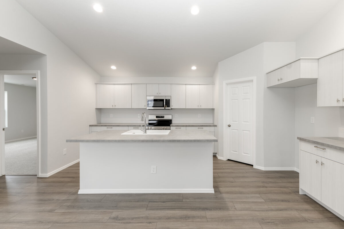 Open-concept kitchen with light gray cabinets, white backsplash, and solid gray countertops. Features a central island with a white integrated sink and brushed nickel faucet. Stainless steel appliances including a microwave and range are built into the cabinetry. The space is well-lit with recessed ceiling lights and warm wood-look flooring throughout.