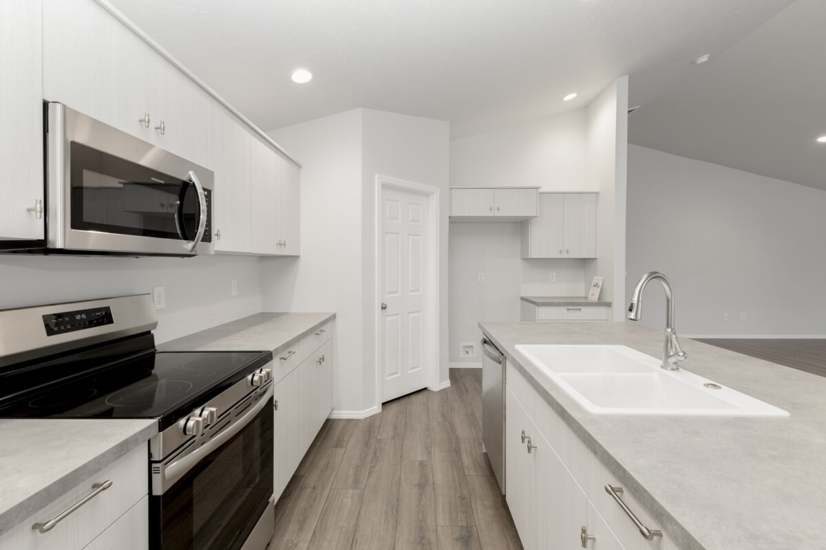 Angled view of a modern kitchen showcasing soft gray cabinets and countertops, a large island with a double-basin white sink, and brushed nickel hardware. Stainless steel microwave and oven are built-in, complemented by vinyl wood plank flooring and a spacious layout open to the main living area.