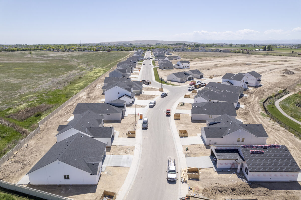 Straight aerial view along the main road of a developing neighborhood, featuring partially completed homes, construction equipment, and green spaces on the left.