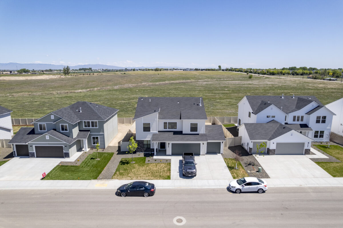 Centered aerial view of three newly built two-story homes with concrete driveways and grass yards, backing up to an open field.