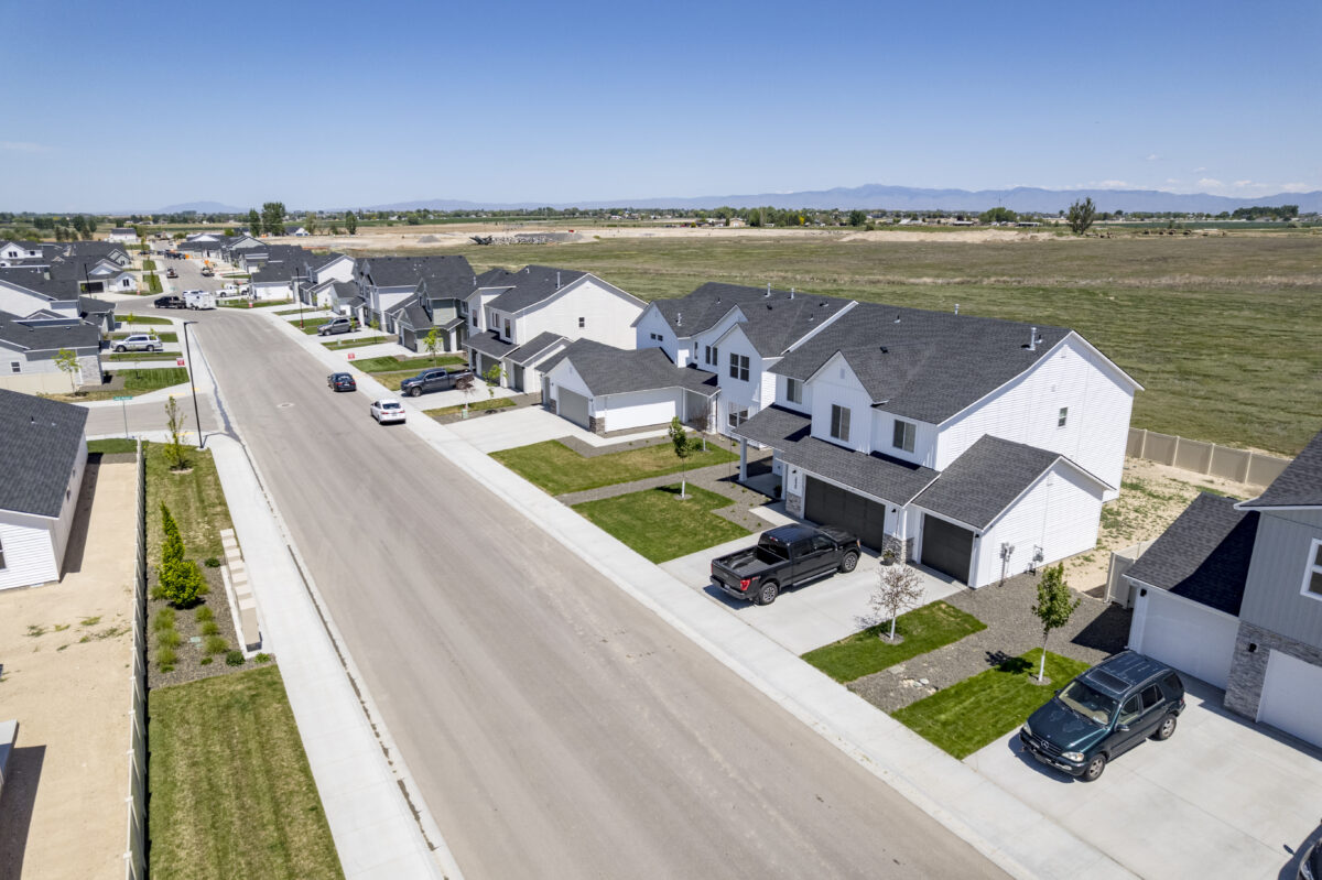 Aerial shot showcasing two-story and single-story homes with dark roofs, neat driveways, and freshly planted yards, running parallel to a large open field.