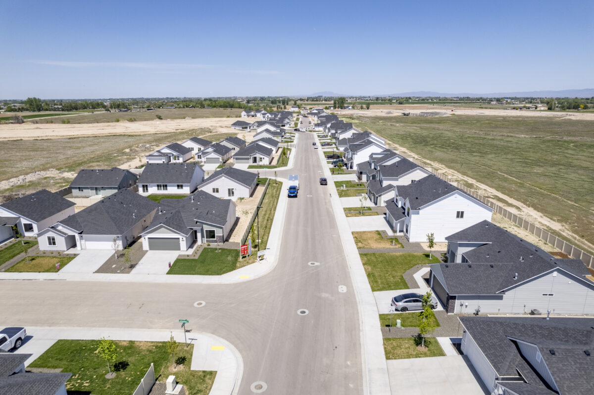 Overhead view down a long residential street lined with identical single-level homes, set beside large undeveloped fields under a bright blue sky.