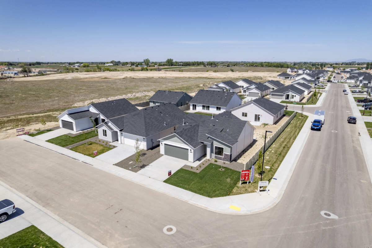 Aerial image of single-story homes on a neighborhood corner lot with freshly laid sod, red CBH Homes signage, and wide streets, surrounded by open fields and clear skies.