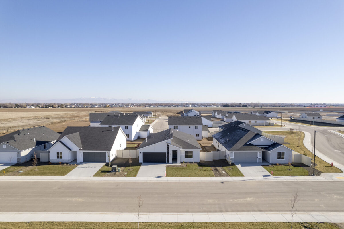 Aerial view of a residential neighborhood featuring modern single-family homes with dark roofs, white exteriors, and neatly landscaped yards, set against a backdrop of open fields and distant mountains under a clear blue sky.