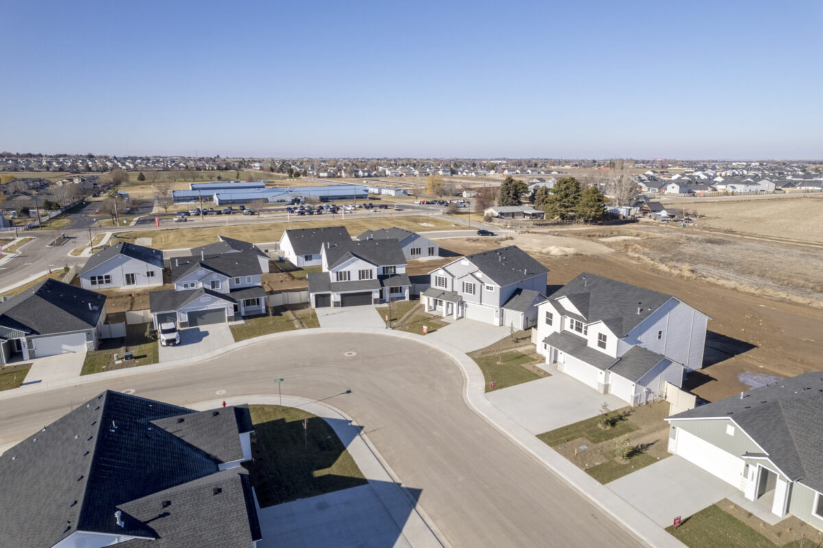 Aerial view of CBH Homes Guches Place community in Caldwell, Idaho with modern two-story homes, dark roofs, and light-colored exteriors, located near open fields, local amenities, and distant residential areas under a clear blue sky.