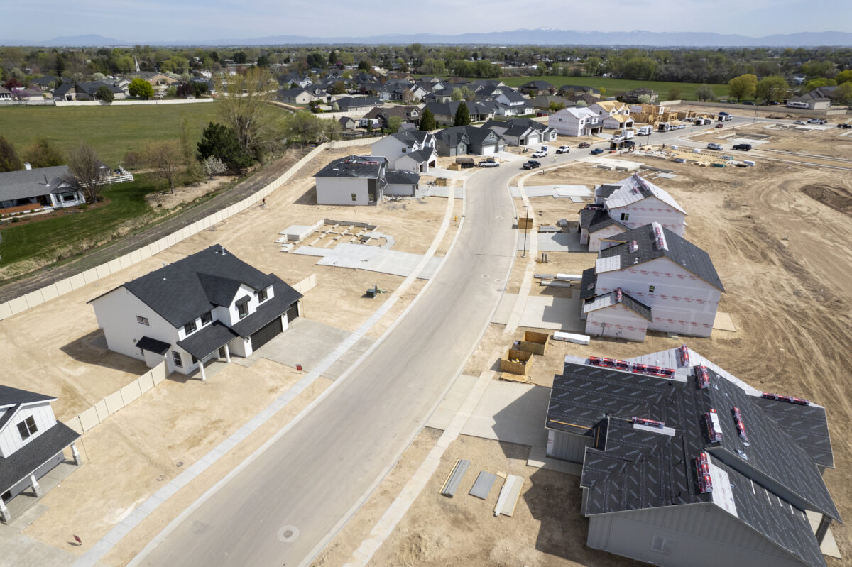 Aerial view of a residential street in a housing development under construction, showing various stages of homebuilding. On the left, completed two-story houses with dark roofs and white exteriors face the road, while on the right, single-story homes are mid-construction, with exposed sheathing and roofing materials stacked on top. Several home foundations are visible in the background, along with construction materials and equipment scattered throughout the site. The development is surrounded by established neighborhoods and green fields, with mountains visible on the distant horizon.
