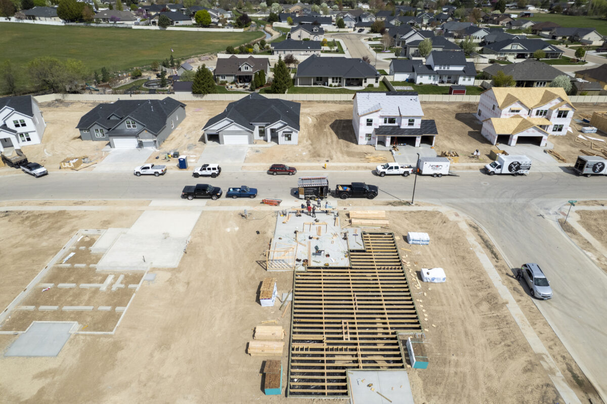 "Overhead view of an active construction site within a residential neighborhood, featuring a framed foundation in the foreground with workers assembling the wooden structure. Across the street, several completed homes and others in various stages of construction—ranging from framed structures to partially roofed buildings—are visible. Construction vehicles and trucks line the street, with neighborhoods and greenery extending into the distance under a clear sky.