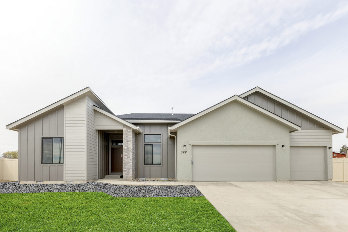 Front view of a modern single-story home with a three-car garage, light gray stucco and siding exterior, and a covered entryway accented by a stone column. The home features clean architectural lines, dark-trimmed windows, and a manicured green lawn bordered by a low rock retaining wall. The house number '5221' is displayed near the garage.