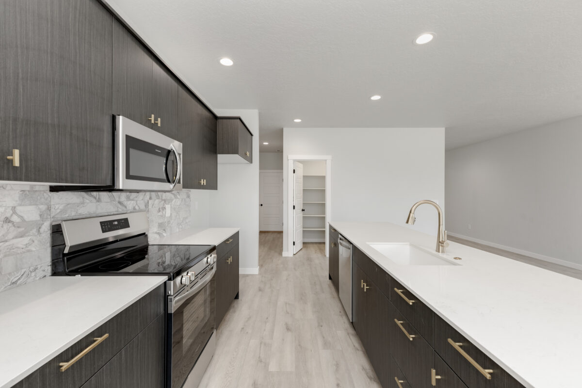 Contemporary kitchen with dark wood cabinetry, gold hardware, and white quartz countertops. A stainless steel oven, microwave, and dishwasher are built into the layout, complemented by a marbled tile backsplash. The kitchen features a large island with an undermount sink and sleek gooseneck faucet, opening toward a spacious living area with light wood-look flooring and neutral walls. A walk-in pantry with white shelving is visible through an open door.