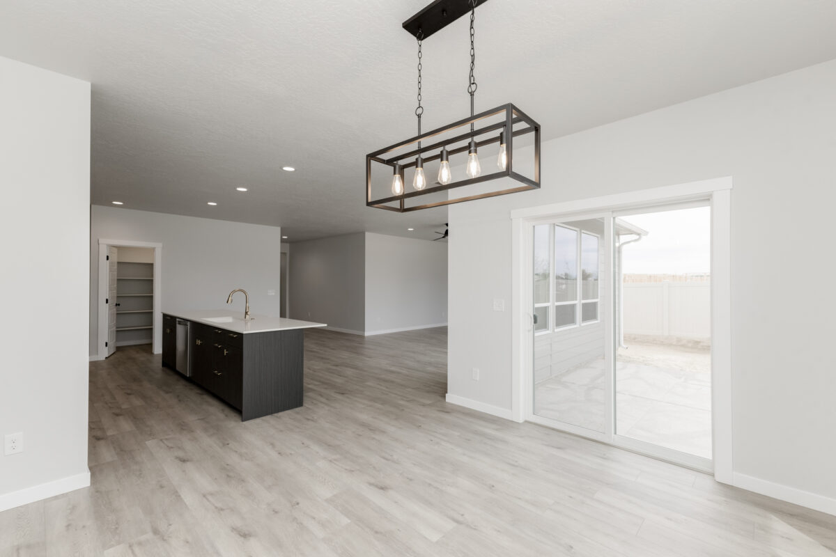 Bright, modern open-concept interior of a new home, showcasing a kitchen with a dark central island, white quartz countertops, stainless steel fixtures, and a walk-in pantry. The space features light wood-style flooring, recessed lighting, and a contemporary black metal chandelier above the dining area. Large sliding glass doors open to a fenced backyard with a concrete patio.