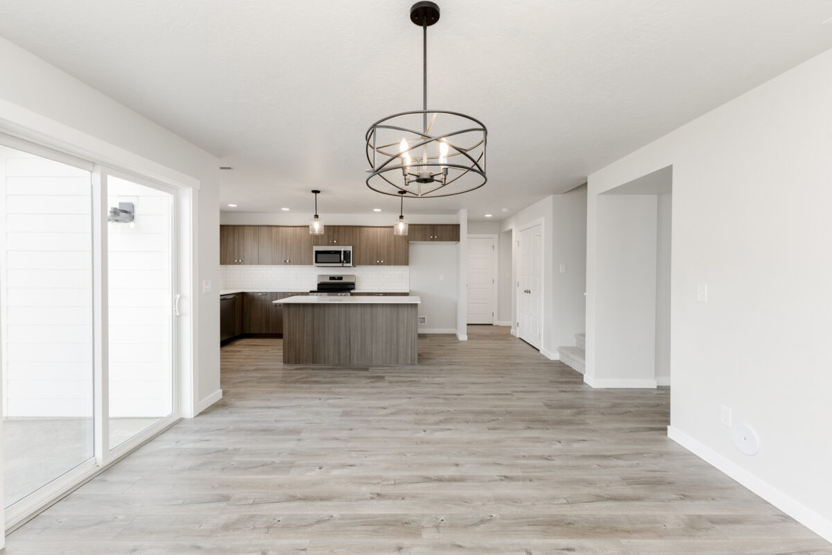 Modern open-concept kitchen and dining area with light wood-style flooring and soft gray walls. The kitchen features a central island with pendant lighting, sleek flat-panel wood cabinets, stainless steel appliances, and a white subway tile backsplash. A round black chandelier hangs over the dining space, which is adjacent to sliding glass doors leading to the backyard.
