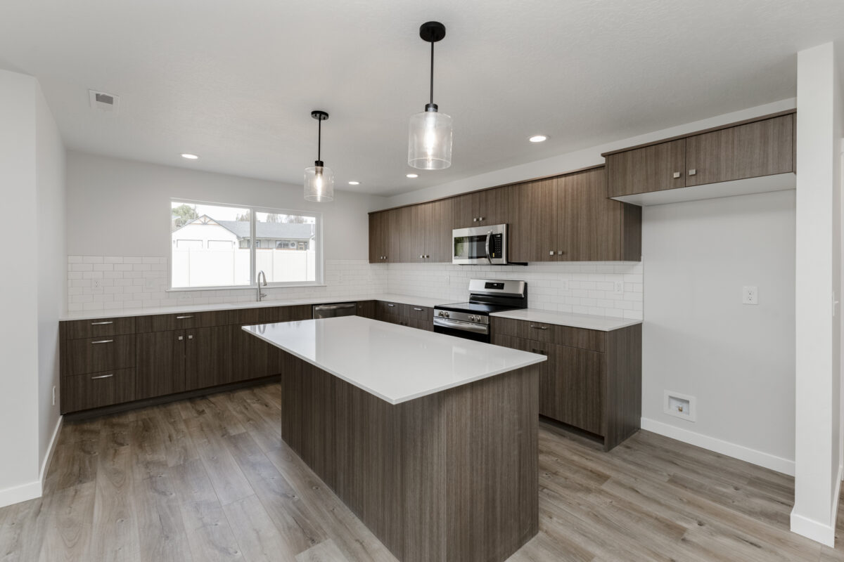"Modern kitchen with wood-grain cabinetry, sleek white quartz countertops, and a large central island. Stainless steel appliances including a microwave, oven, and dishwasher are built in, complemented by a white subway tile backsplash. Two clear glass pendant lights hang above the island, and natural light enters through a wide window over the sink. The flooring is a light-toned wood-style laminate, and an open space to the right appears ready for a refrigerator.
