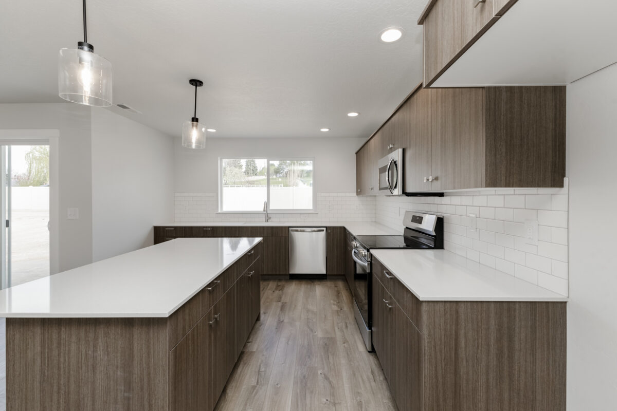 Contemporary kitchen with dark wood cabinetry, gold hardware, and white quartz countertops. A stainless steel oven, microwave, and dishwasher are built into the layout, complemented by a marbled tile backsplash. The kitchen features a large island with an undermount sink and sleek gooseneck faucet, opening toward a spacious living area with light wood-look flooring and neutral walls. A walk-in pantry with white shelving is visible through an open door.