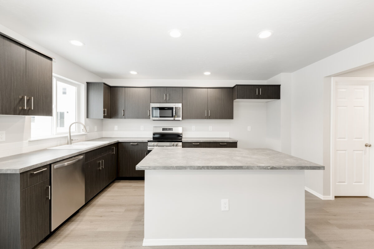 Modern kitchen design with espresso-tone cabinets and contrasting gray concrete-style countertops. Features a large island, white backsplash, stainless steel appliances including a microwave and dishwasher, and under-cabinet lighting. The space is brightened by recessed ceiling lights and a large window over the sink, all set against a light-toned wood-look floor.