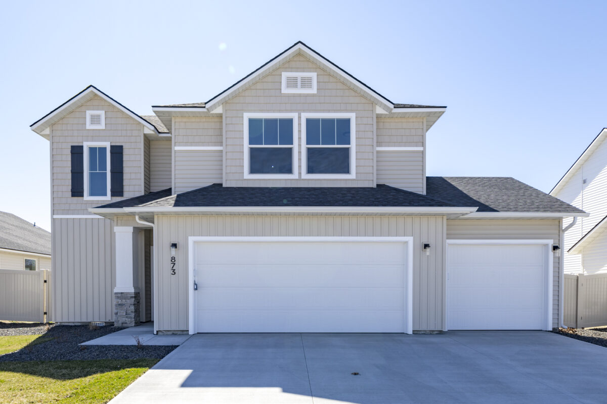 Stylish two-story beige suburban home with white trim and varied textures including vertical and shingle-style siding. The house includes a three-car garage with white doors, a covered entry with a white post and stone column, and decorative black shutters on one window. The driveway and lawn are well-maintained, giving a clean, welcoming appearance.