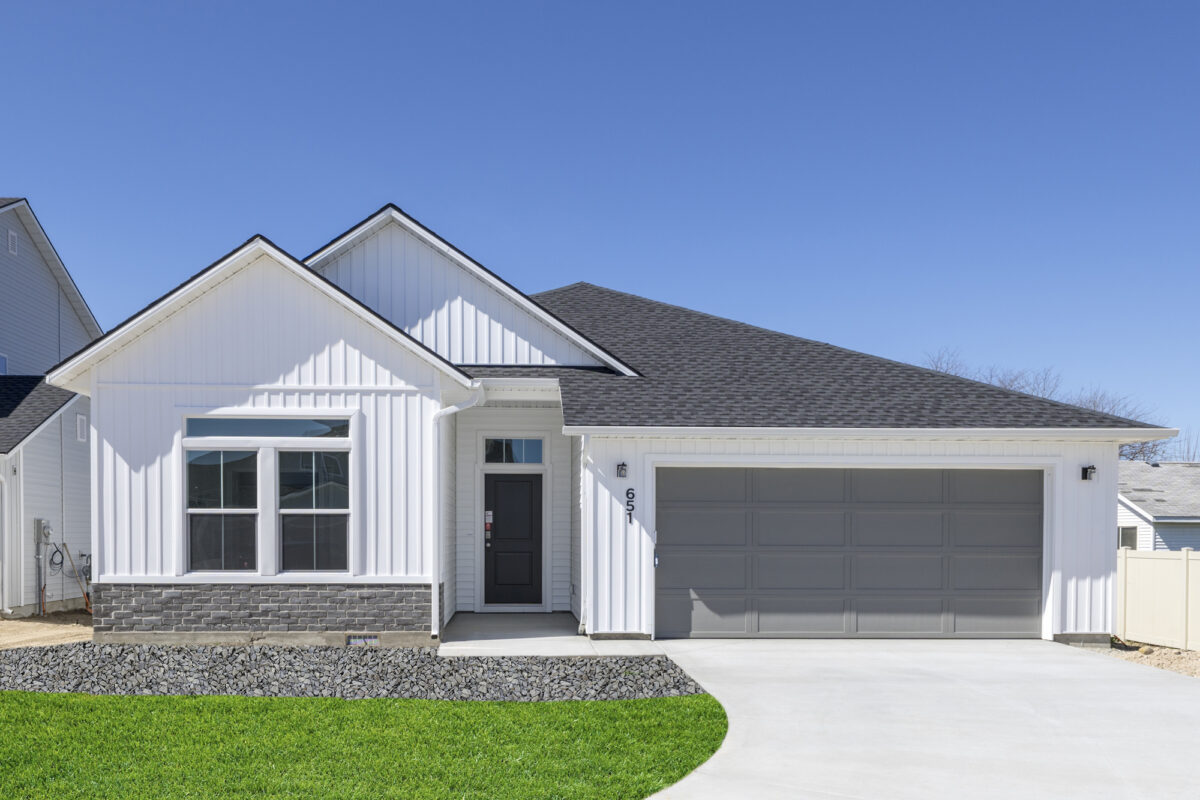 Front elevation of a single-story home with crisp white board-and-batten siding, dark gray two-car garage, and black front door. The home features stone wainscoting beneath the front window and is surrounded by neatly landscaped green grass and decorative rock accents under a clear blue sky.