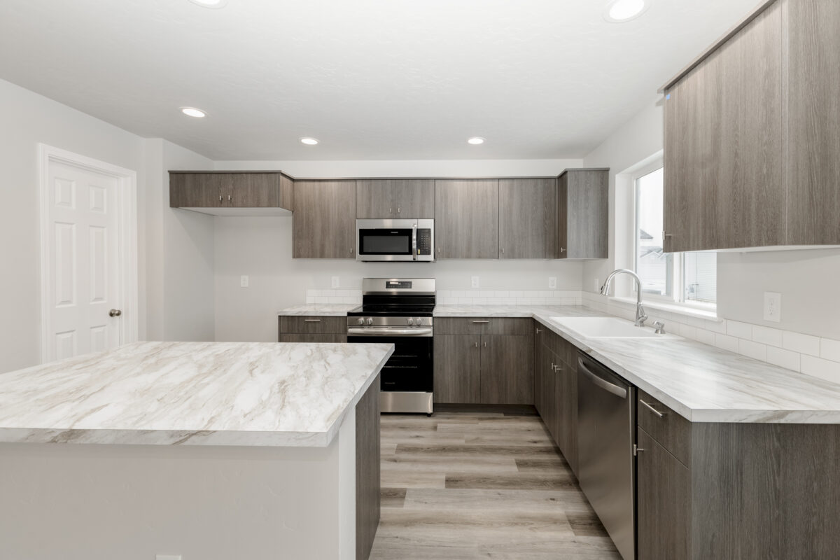 Contemporary kitchen with dark wood-tone cabinets and sleek brushed nickel hardware. The space includes white marble-look countertops, a center island, and stainless steel appliances. Natural light streams in through a large window above the sink, complementing the clean white tile backsplash and neutral-toned vinyl plank flooring.