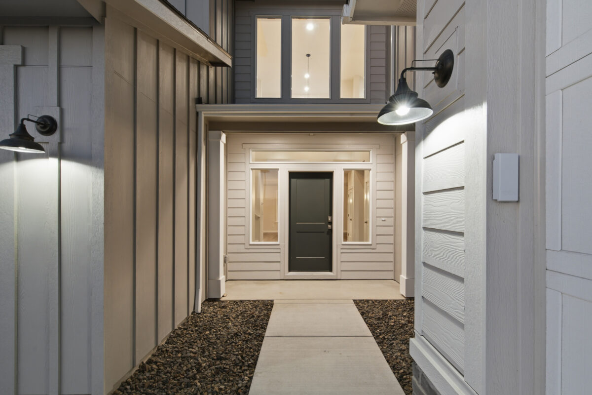 Modern two-story home entrance at dusk featuring a covered walkway, black front door, illuminated exterior sconces, and a mix of vertical board-and-batten and horizontal lap siding in soft gray tones.