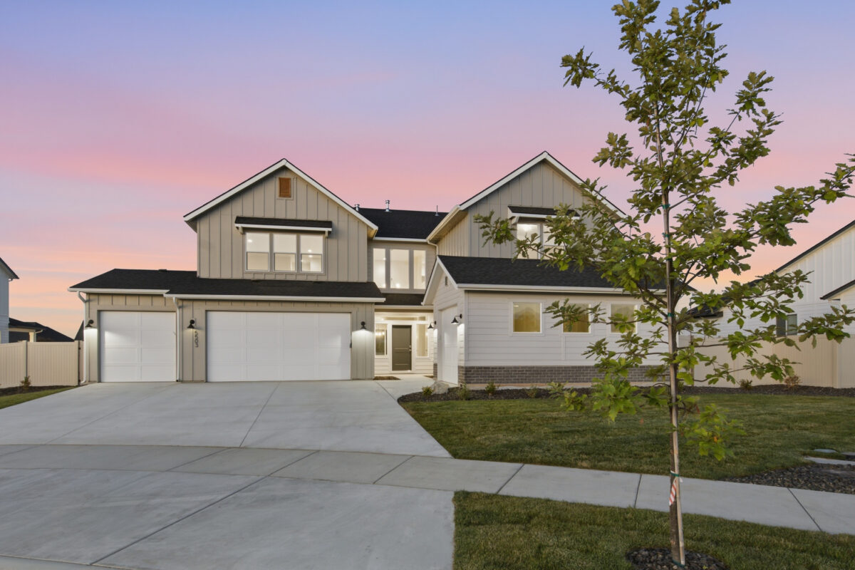 Two-story modern farmhouse-style home at twilight featuring light gray board-and-batten siding, a three-car garage, landscaped front yard, and warm exterior lighting beneath a pink and blue sunset sky.