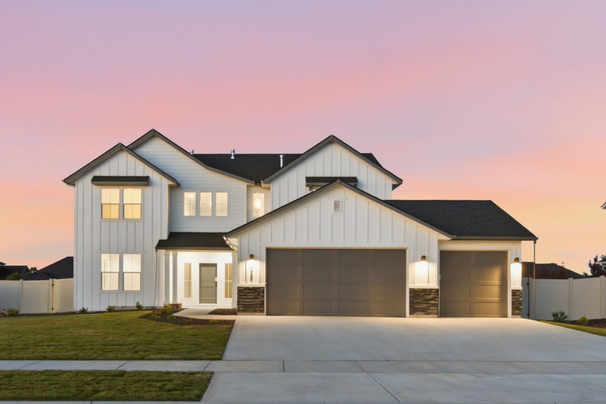 Two-story modern farmhouse-style home at sunset featuring white board-and-batten siding, a three-car garage, stone accents, and warm exterior lighting beneath a pink evening sky.