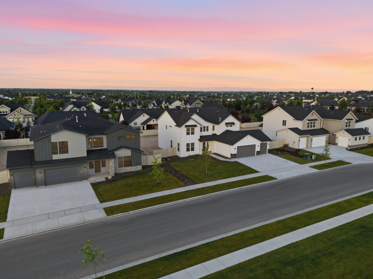 Aerial view of a new residential neighborhood at sunset featuring modern homes, landscaped yards, wide streets, and colorful pink and orange skies stretching across the horizon.