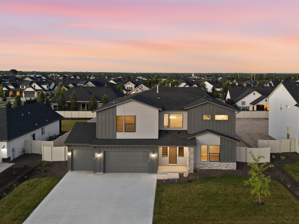 Aerial twilight view of a modern two-story home with gray and white siding, a three-car garage, landscaped yard, and warm interior lighting in a new residential community.