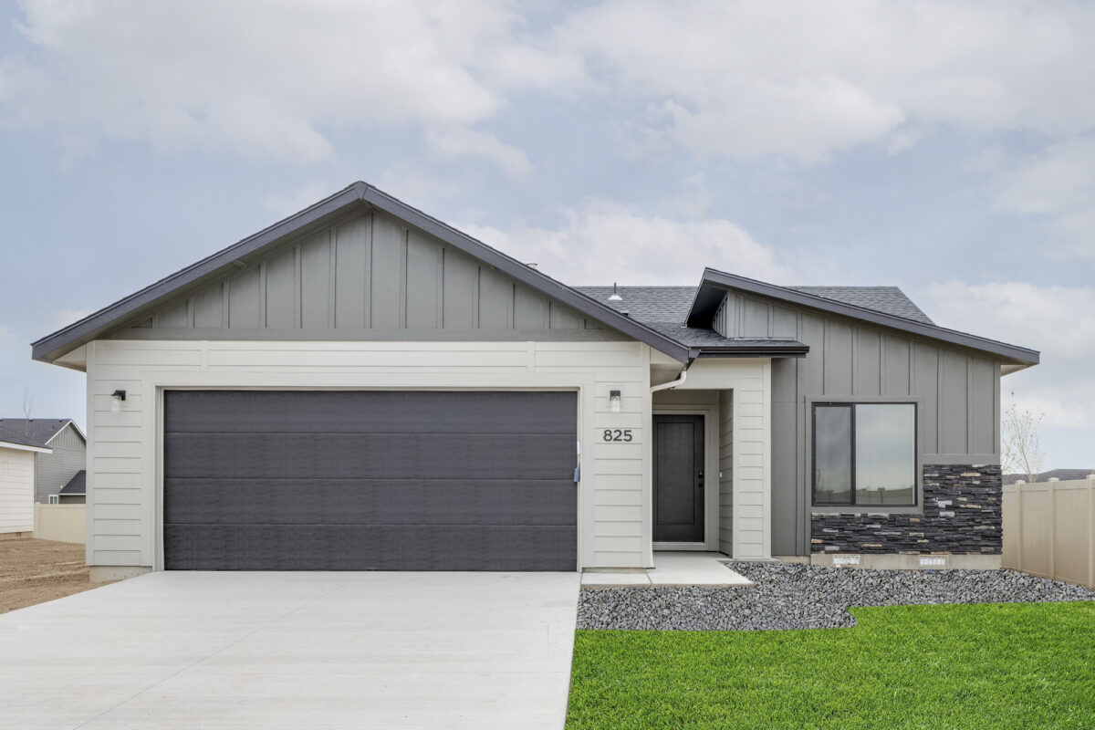 Contemporary ranch-style home with a two-tone exterior—light siding below and dark gables above—paired with a bold black garage door. Includes modern stone accents around a wide window, vertical board-and-batten siding, and a covered front entryway. Crisp landscaping with decorative rock and lush green lawn complete the look.