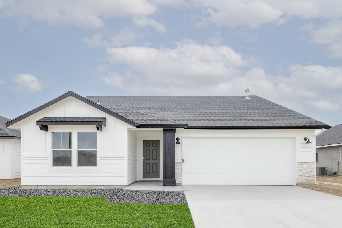 Modern single-story home with crisp white board-and-batten siding, a black accent column and front door, and a white two-car garage. Features a decorative front window awning, black trim, and rock landscaping with fresh green grass beneath a soft, cloud-dappled sky.