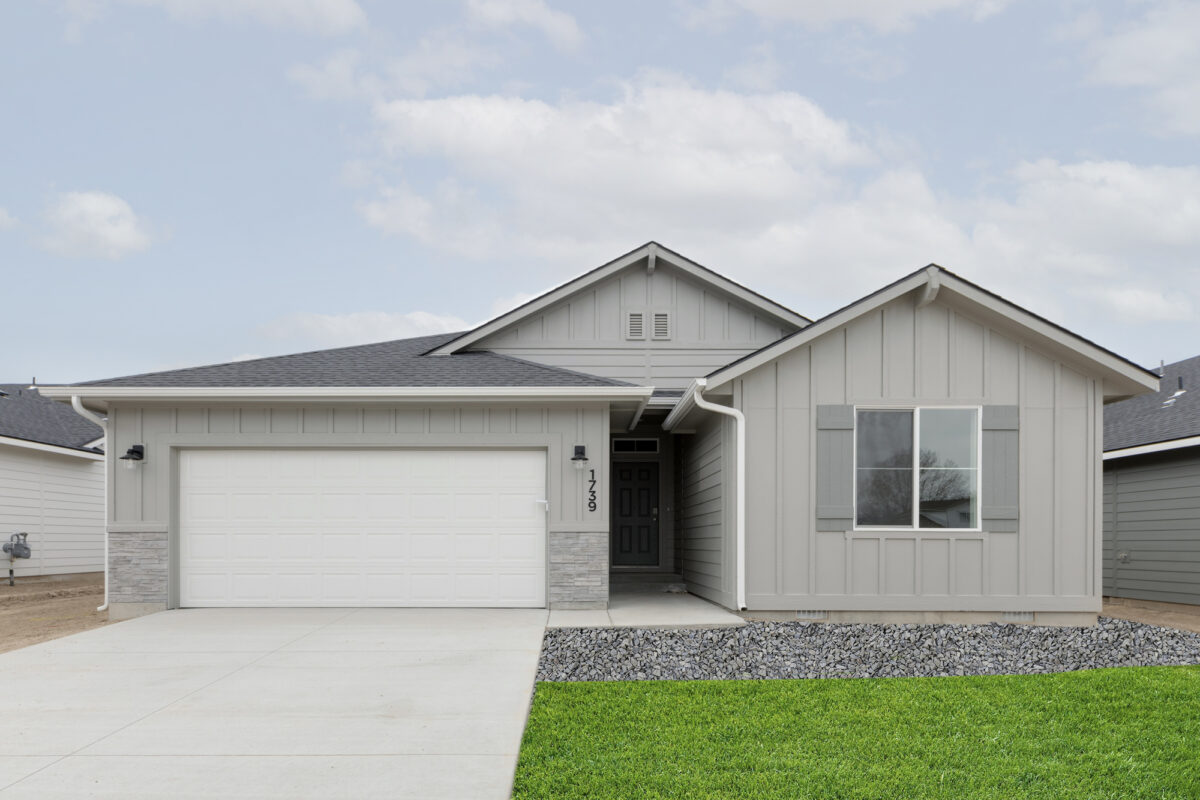 Clean and balanced single-story gray home with vertical board-and-batten siding, light stone accents, and a white garage door. The entrance is recessed and framed with a dark front door. Features shutters flanking the front window and a curved rock bed leading into the concrete driveway.