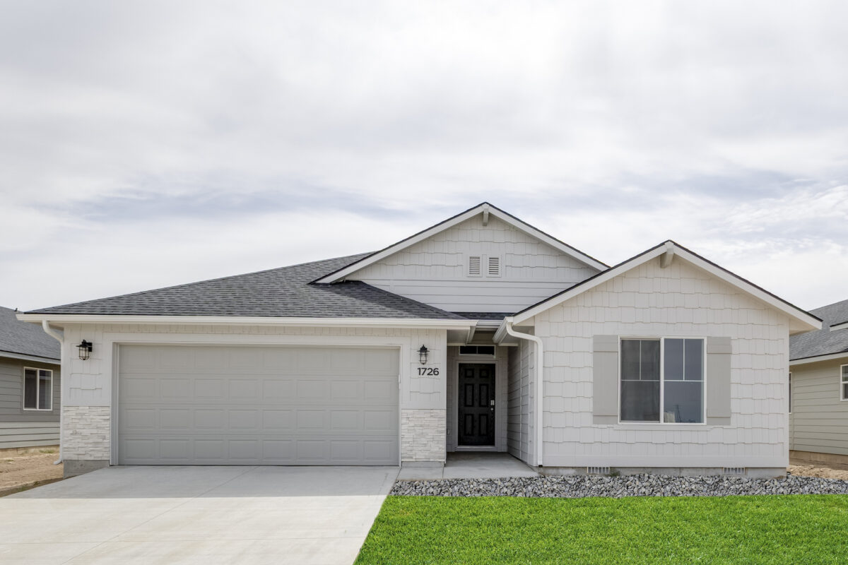 Charming one-story home with a shingle-style gable facade, white and gray siding, and white stacked stone accents. The home includes a soft gray garage door, dark front door, and tidy landscaping featuring rock beds and a healthy green lawn.