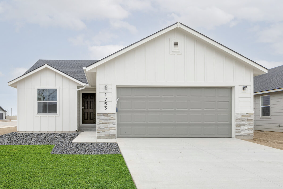 Bright and simple single-story home with white vertical siding, light stone base trim, and a soft gray garage door. A covered entry is framed by white columns and leads to a black front door. Minimalist landscaping features dark decorative rock and a clean concrete driveway.