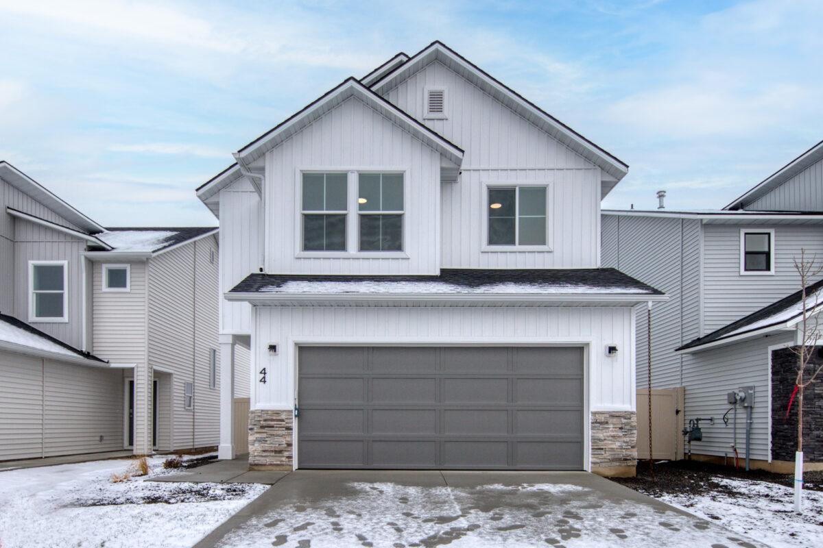 Front view of a two-story modern suburban home with white vertical siding, a gray two-car garage door, and stone accents along the lower exterior. Light snow covers parts of the roof, lawn, and driveway, indicating a winter setting. The house is flanked by similar neighboring homes under a partly cloudy sky.