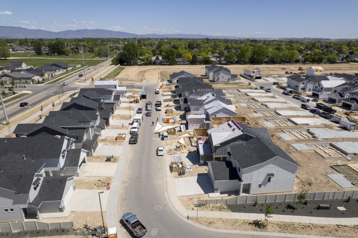 Drone view of an active home construction site showing partially framed houses, finished homes to the left, and cleared foundations ready for vertical build, along a curving residential street.